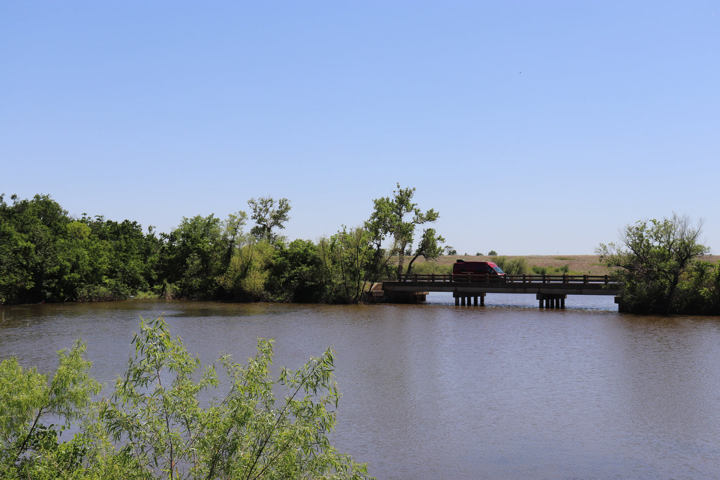 Red RV rental crosses a bridge over a river, surrounded by green nature under a clear sky.
