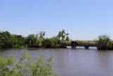 Red RV rental crosses a bridge over a river, surrounded by green nature under a clear sky.