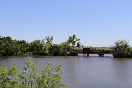 Red RV rental crosses a bridge over a river, surrounded by green nature under a clear sky.