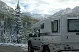 A white RV rental camper truck on a snowy mountain road, surrounded by snow-dusted pine trees under cloudy skies.