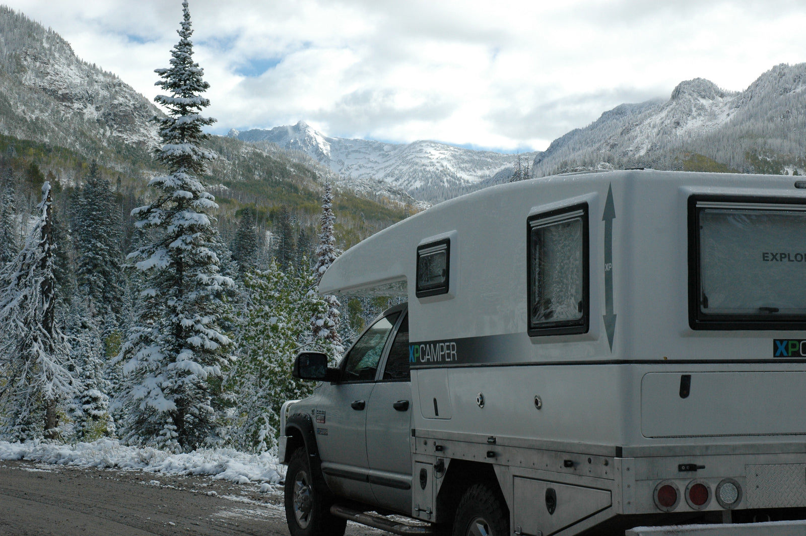 A white RV rental camper truck on a snowy mountain road, surrounded by snow-dusted pine trees under cloudy skies.