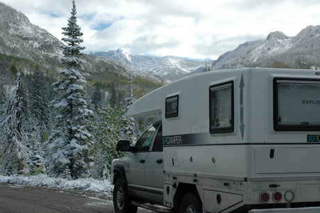 A white RV rental camper truck on a snowy mountain road, surrounded by snow-dusted pine trees under cloudy skies.