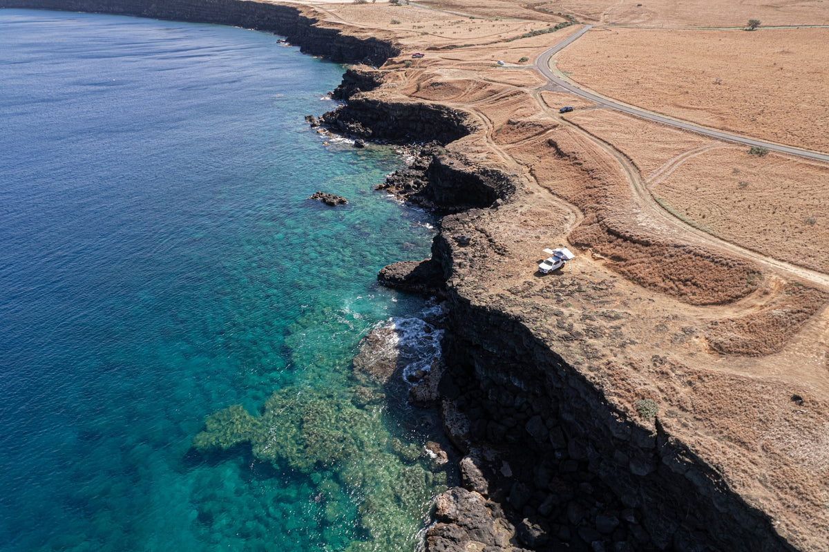Aerial view: A white RV rental truck with a rooftop tent on a dry cliff overlooking the clear blue ocean. — photo 9