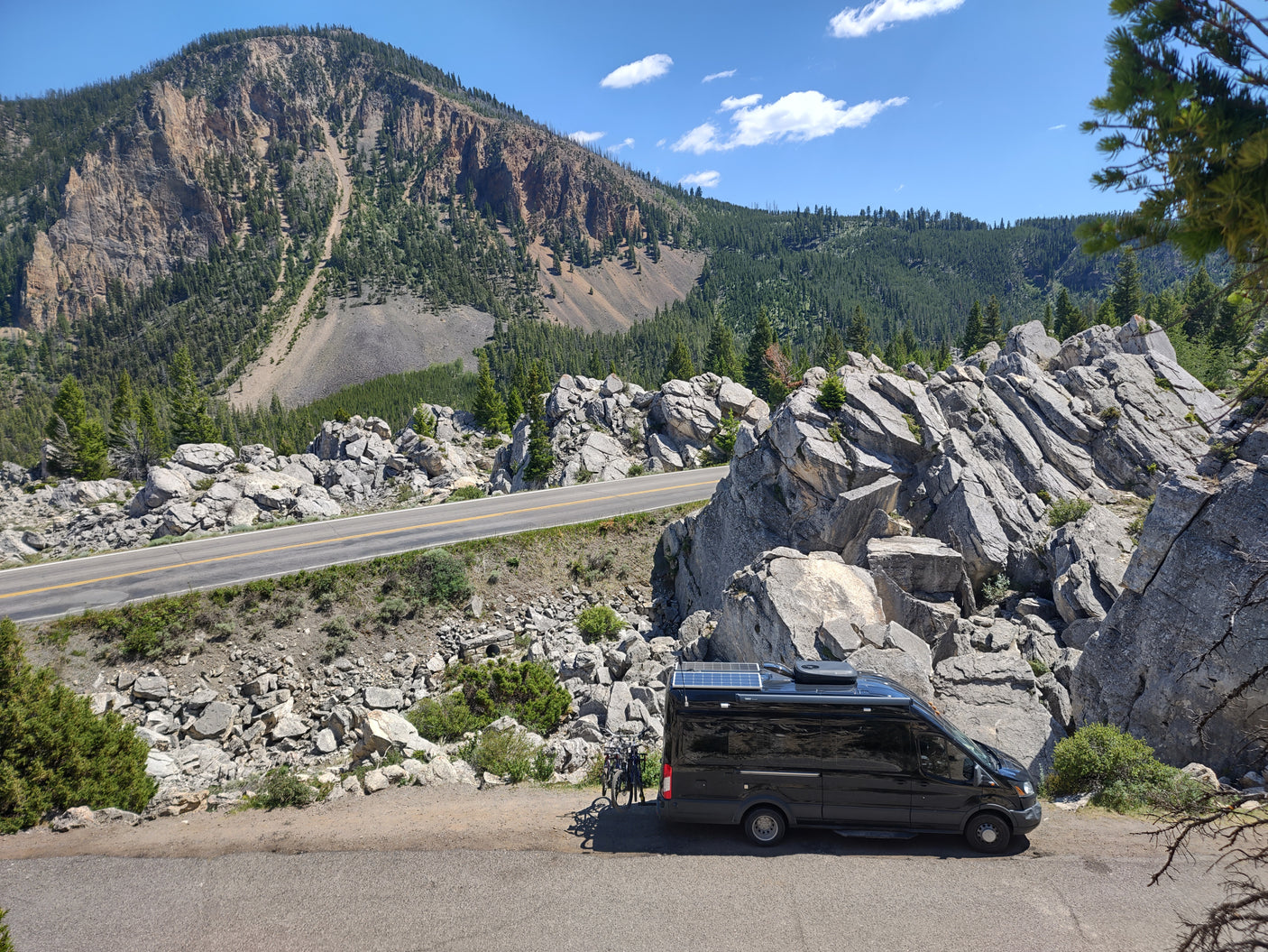 An RV rental camper van with bikes, parked on a scenic winding mountain road, amidst rocky cliffs and green forest.