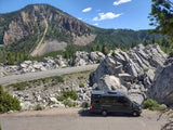 An RV rental camper van with bikes, parked on a scenic winding mountain road, amidst rocky cliffs and green forest.