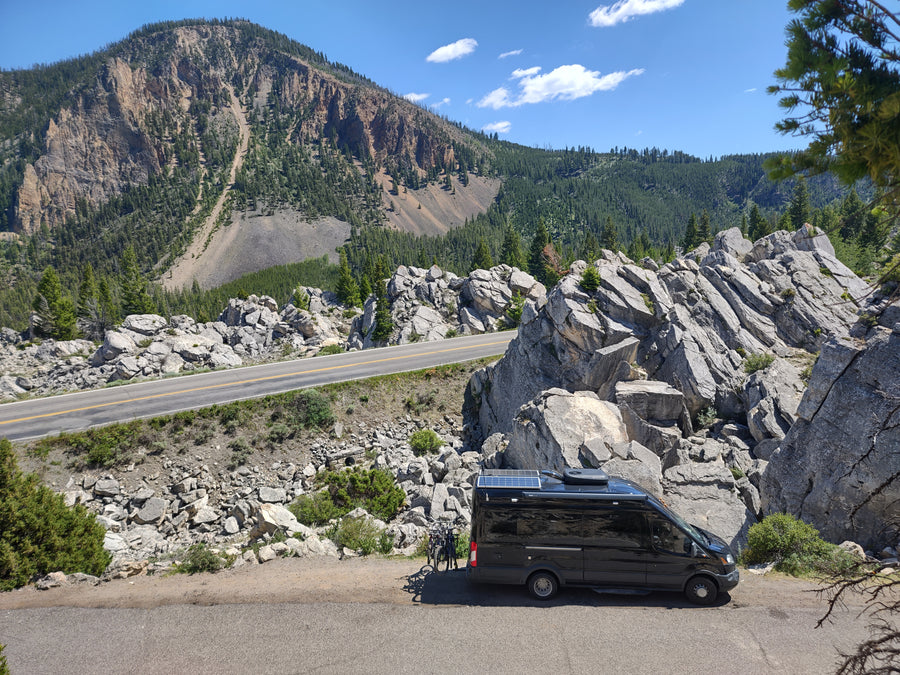 An RV rental camper van with bikes, parked on a scenic winding mountain road, amidst rocky cliffs and green forest.