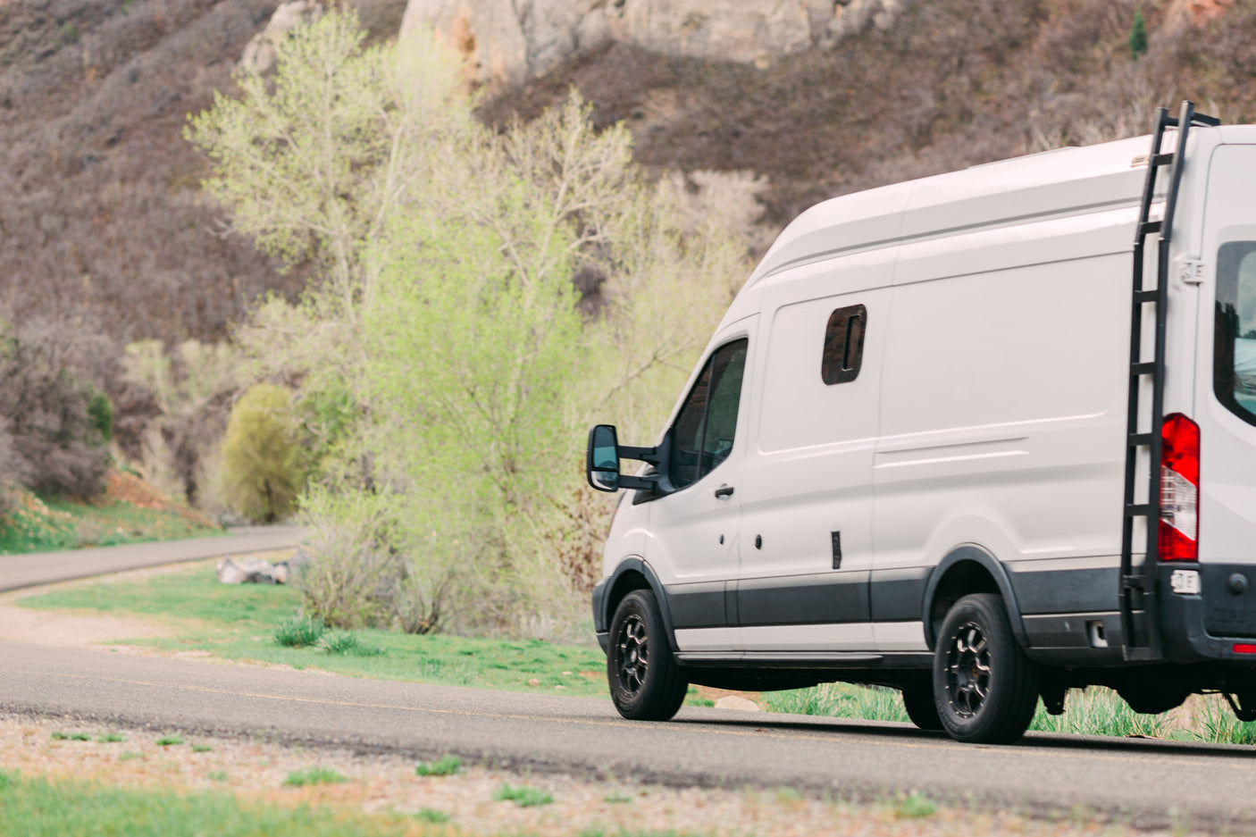 A white RV rental campervan with a rear ladder travels on a scenic road amidst spring foliage.
