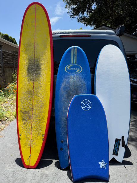 Colorful surfboards & a bodyboard lean on an RV rental van, likely for a beach trip. Sun shines on boards in the driveway.