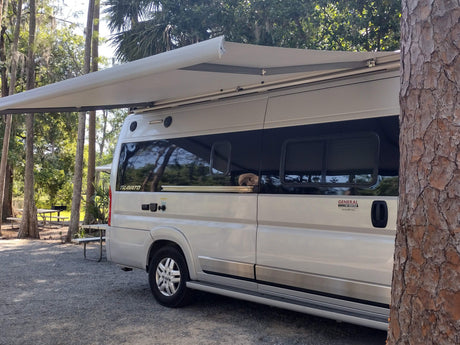 An rv rental camper van, with an extended awning, is nestled in a shaded campsite. A dog peeks from a window, enjoying the view.