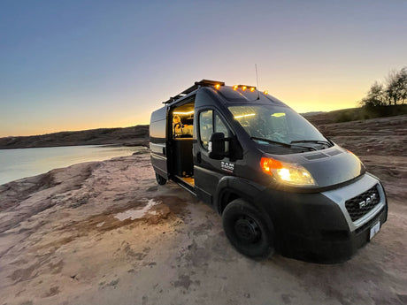 Scenic rv rental parked by the water at sunset. Interior lights glow, showing a person inside this adventure-ready campervan.