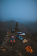Four SUVs & two tents perched on a misty mountain cliff, overlooking forests. Rugged off-road camping is a unique adventure, unlike an RV rental.