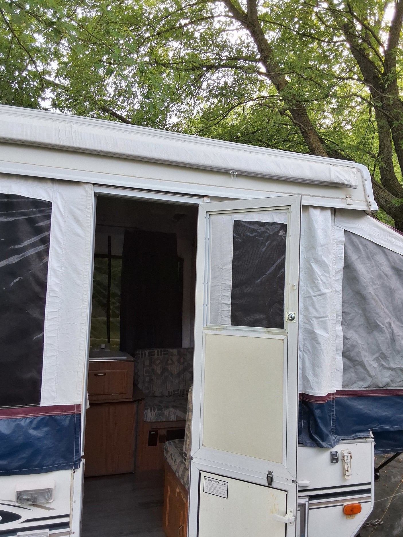 Pop-up rv rental camper with its door open, showing a compact interior with a sink, cabinet, and seating, nestled among green trees.