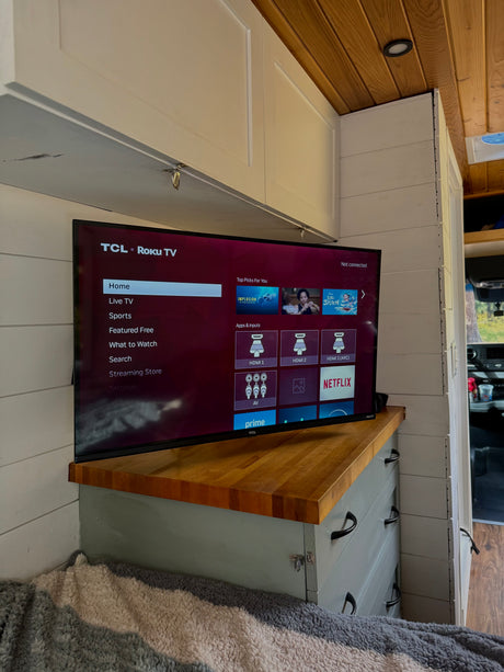 This RV rental features a TCL Roku TV displaying streaming apps and inputs on a wooden counter, part of a cozy custom-built interior.