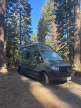 A dark green Sprinter RV rental is parked on a dirt path in a sunny forest campground among tall trees.
