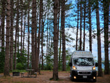 White RV rental parked among tall pine trees at a campsite with a picnic table and fire pit, overlooking distant water.