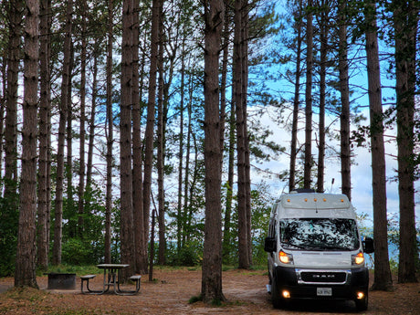 White RV rental parked among tall pine trees at a campsite with a picnic table and fire pit, overlooking distant water.