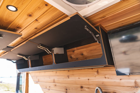 Inside an RV rental: custom wood paneling, black overhead cabinets with lift-up doors, integrated LED lighting, and a TV for entertainment.