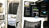 RV rental interior: A camper van's control panel, showing switches and displays for heating, water, and solar charge, beside a cozy bed.