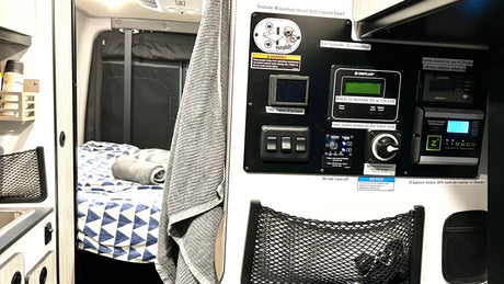 RV rental interior: A camper van's control panel, showing switches and displays for heating, water, and solar charge, beside a cozy bed.