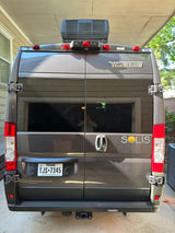 Winnebago Solis rv rental: rear view of a grey camper van with rooftop AC & Texas plate, parked under a carport.