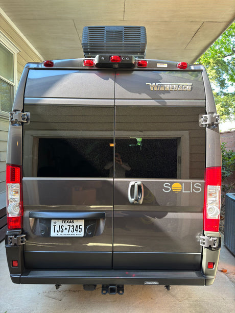 Winnebago Solis rv rental: rear view of a grey camper van with rooftop AC & Texas plate, parked under a carport.