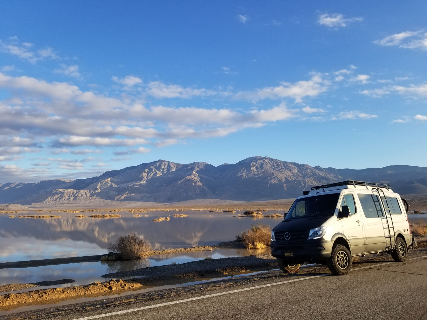 A white adventure rv rental van, likely a Sprinter, is parked by a reflective lake in a mountainous desert. Ideal for road trips!