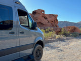 Adventure awaits! This gray RV rental van is parked on a gravel path, showcasing red rock formations under a clear sky.