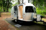 A sleek silver Airstream rv rental awaits in a serene forest. Its door is open, inviting guests for a unique camping experience.