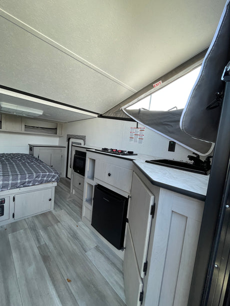 Bright rv rental interior featuring a bed, kitchenette with stove, sink, microwave, mini-fridge, and pop-up roof. Light wood finishes.