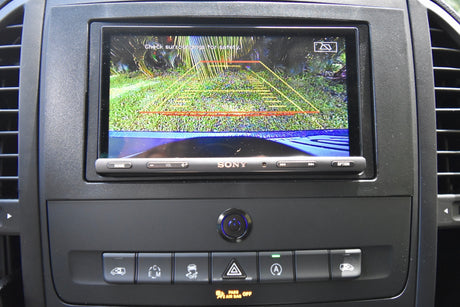 Dashboard of an RV rental with a clear backup camera view displaying safety lines and surrounding environment. Various control buttons are visible below.