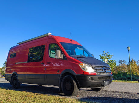 Red and black Mercedes-Benz Sprinter camper van, equipped with a roof awning, perfect for an RV rental getaway.