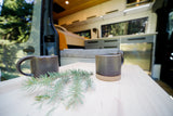 Enjoy nature from your RV rental! Two ceramic mugs and evergreen sprigs on a wood table inside a modern camper van.