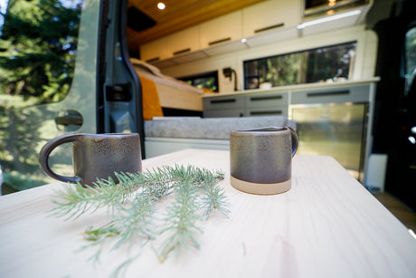 Enjoy nature from your RV rental! Two ceramic mugs and evergreen sprigs on a wood table inside a modern camper van.
