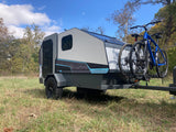 Sleek RV rental camper with two bikes mounted, parked in a sunny, grassy field under a blue sky, perfect for outdoor escapes.