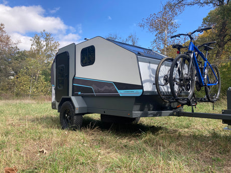 Sleek RV rental camper with two bikes mounted, parked in a sunny, grassy field under a blue sky, perfect for outdoor escapes.