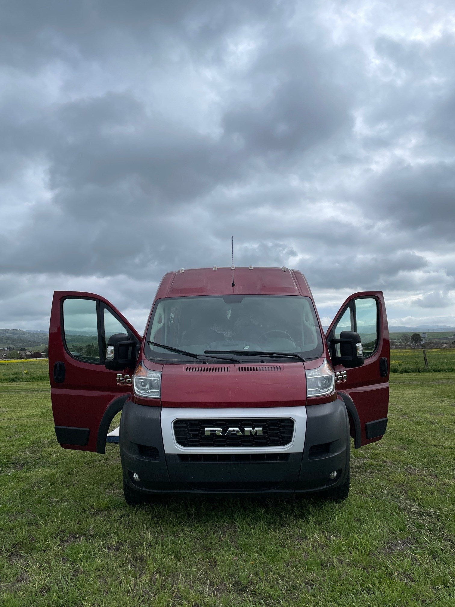 Red Ram ProMaster van for RV rental, doors open in a green field under cloudy skies. Your travel adventure starts here!