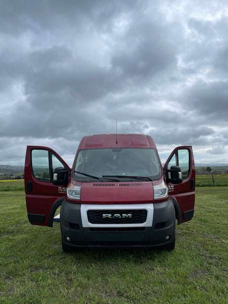 Red Ram ProMaster van for RV rental, doors open in a green field under cloudy skies. Your travel adventure starts here!