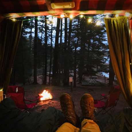Cozy RV rental view: feet up, string lights on, watching the campfire glow by the lake and forest. Perfect camping escape.