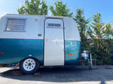 Classic teal & white travel trailer RV rental. Parked outdoors with green trees & blue sky.