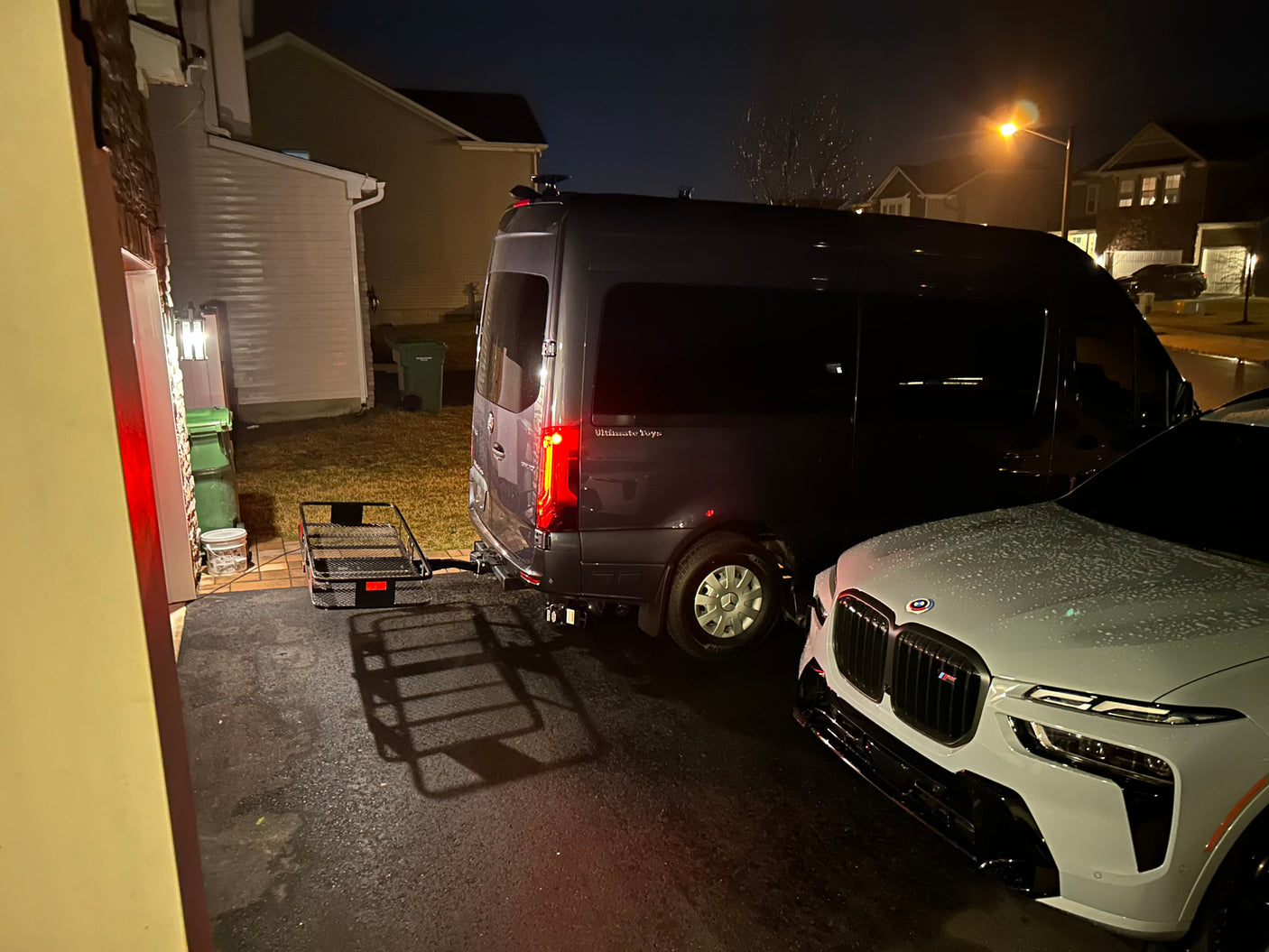 A dark gray luxury RV rental van with a cargo hitch carrier is parked next to a white BMW SUV at night.