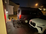 A dark gray luxury RV rental van with a cargo hitch carrier is parked next to a white BMW SUV at night.