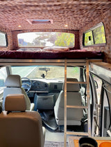 Inside an rv rental, showing a raised bed above the driver's cabin, accessible by a ladder. Unique patterned walls and ceiling.
