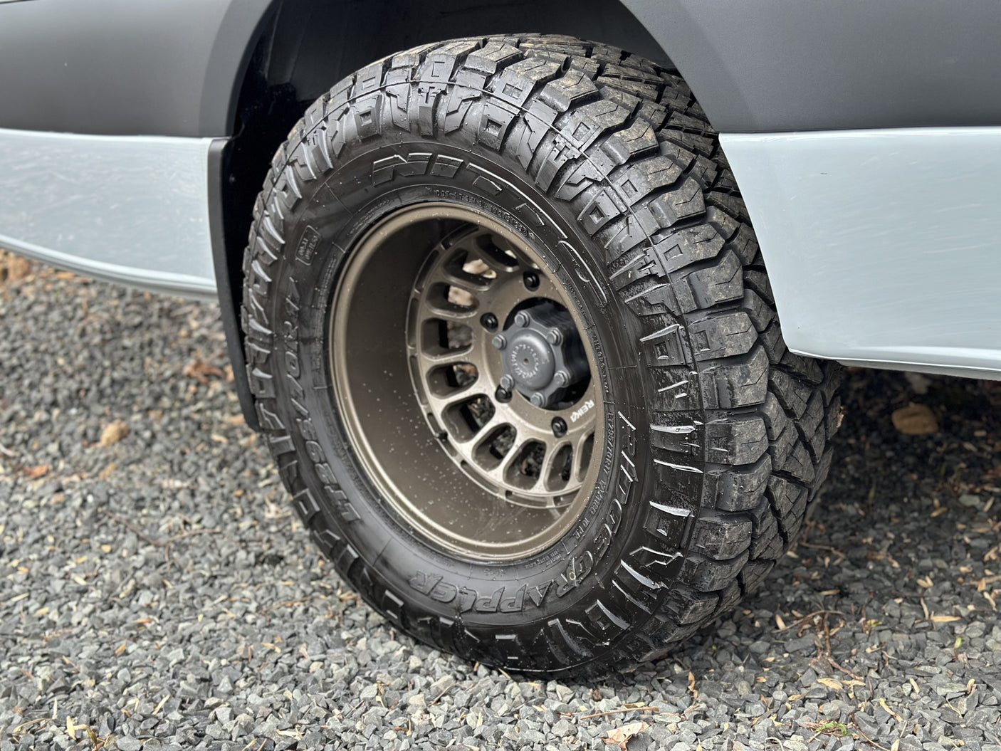 Off-road tire and custom wheel on an adventure-ready RV rental, parked on a gravel surface.