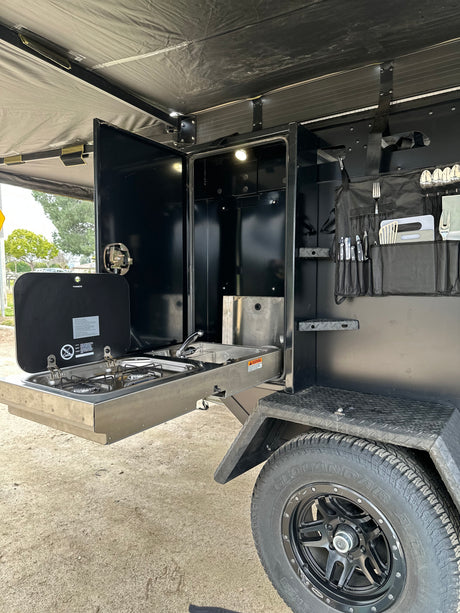 Get ready for camping with this RV rental! Black camper trailer shows its pull-out kitchen: stove, sink, and organized utensils.