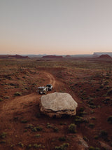 An RV rental off-road setup: truck & trailer on a desert path. A person is next to it, with red mesas & vast sky in the distance.