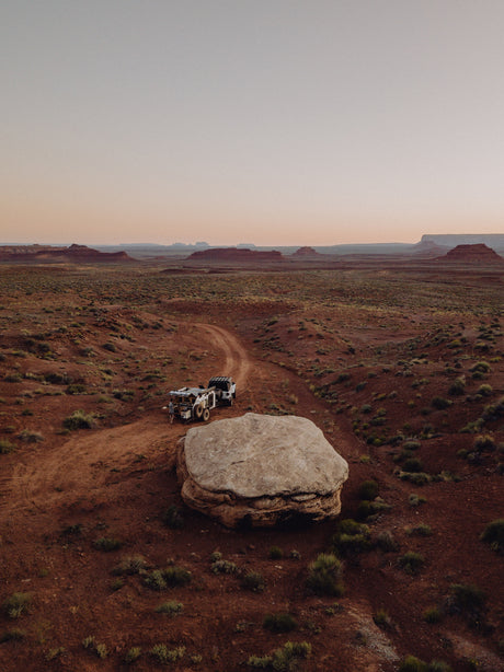 An RV rental off-road setup: truck & trailer on a desert path. A person is next to it, with red mesas & vast sky in the distance.