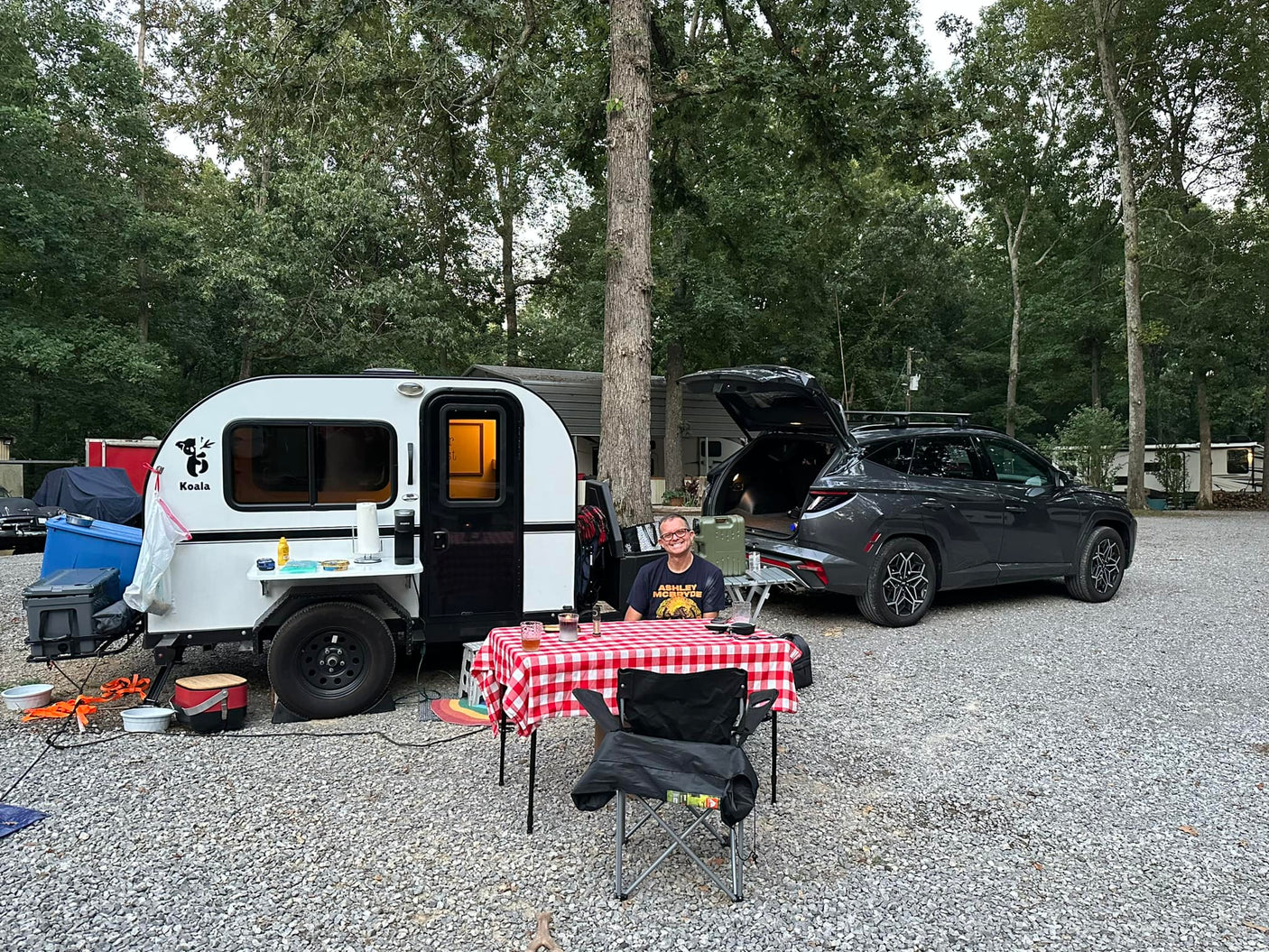 A man is camping with an RV rental Koala teardrop camper and SUV, sitting at a checkered picnic table.