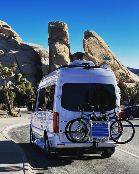 Exploring Joshua Tree: White RV rental with bikes parked amidst majestic rock formations under a clear blue sky.