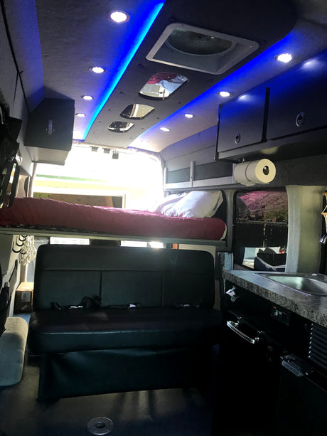 Compact RV rental interior featuring blue LED lighting, a convertible bed, and small kitchen setup for comfortable travel.
