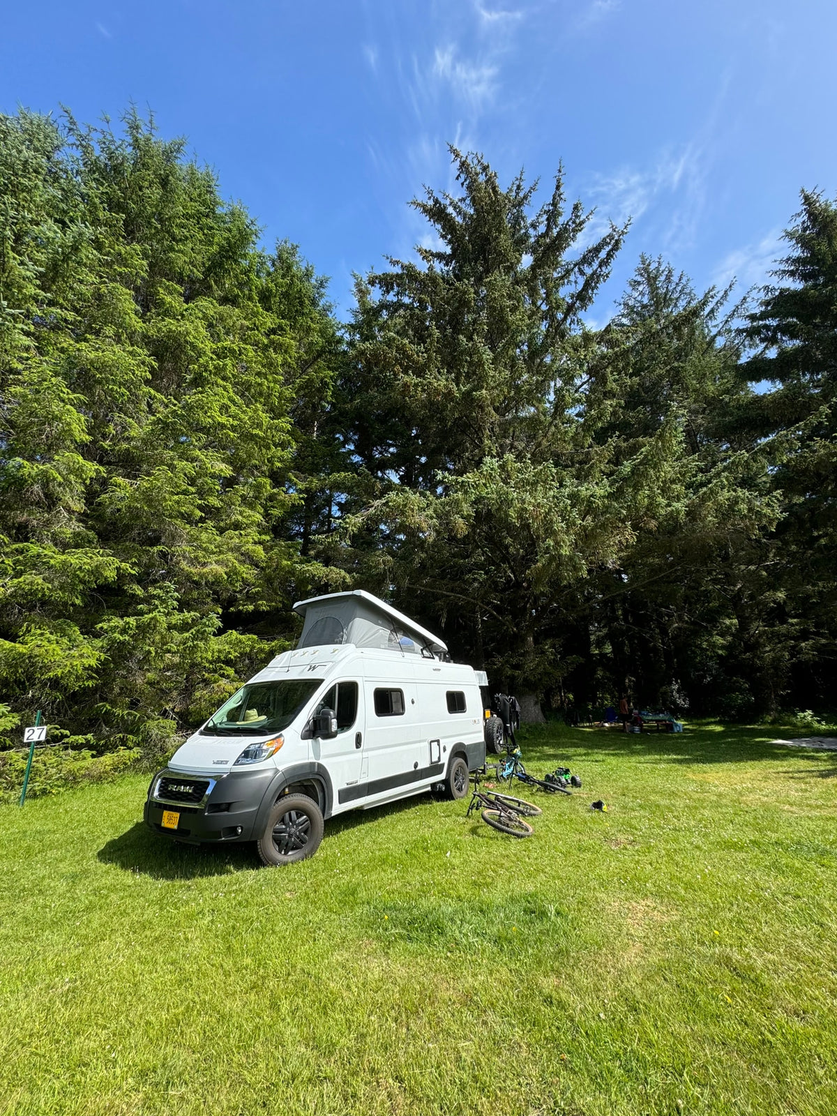 White camper van (RV rental) with pop-top at a green campsite, surrounded by tall trees and blue sky. Bicycles suggest nature exploration.